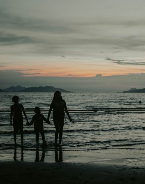Silhouette of three people holding hands along the shore of water at sunset.